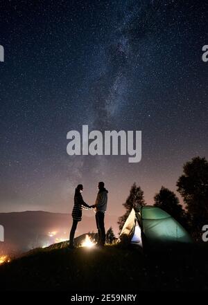 Silhouette de couple romantique heureux touristes debout au feu de camp près de la tente touristique, tenant les mains sous le ciel de nuit plein d'étoiles et de voie lactée. En arrière-plan ciel étoilé, montagnes et ville lumineuse Banque D'Images