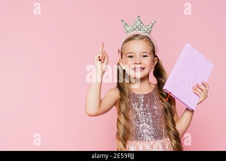 sourire petite fille dans la couronne tenant le livre et pointant avec doigt isolé sur rose Banque D'Images