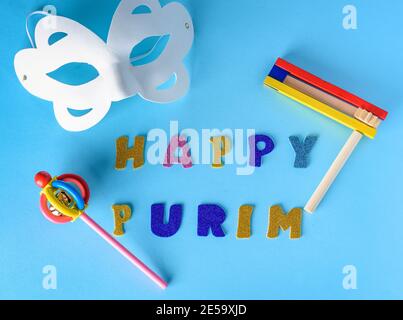 Carte de voeux Happy Purim Jewish Celebration Holiday. Masque de carnaval blanc, bruiteurs en bois colorés ou graggers sur fond bleu.Happy Purim paillettes text.Flat Lay. Banque D'Images