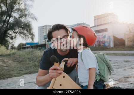 Un père souriant embrasse son jeune fils à vélo. Un petit garçon portant un casque embrassant son papa lors d'une marche en soirée. Banque D'Images