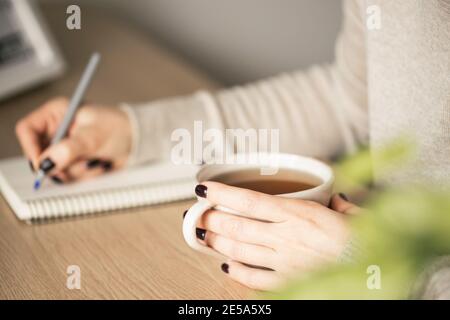 Femme en chandail beige tenant une tasse de téain d'une main et elle écrit. Mise au point sélective. Banque D'Images