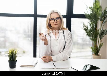 Chat vidéo avec une femme médecin avec un stéthoscope en blouse de laboratoire assis au bureau, portant des lunettes et regardant la caméra, donnant la consultation Banque D'Images