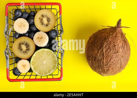 Panier en métal rouge plein de fruits et de baies isolées sur fond jaune. Citron vert, noix de coco, banane, bleuets, kiwi et pommes dans une boîte à provisions. Cocktail de vitamines. . Banque D'Images