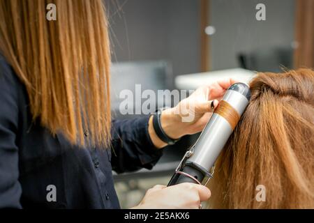 Coiffeur professionnel fait des boucles avec un fer à friser pour un jeune femme aux longs cheveux rouges dans un salon de beauté Banque D'Images