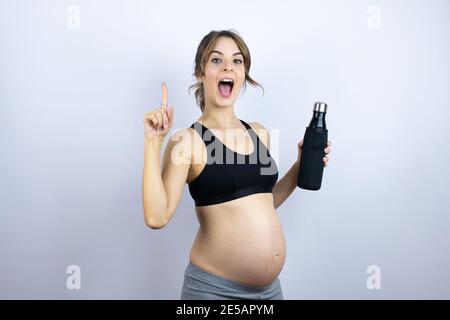 Jeune sportif enceinte portant des vêtements de sport tenant une bouteille avec de l'eau arrière-plan blanc stupéfié et surpris en regardant l'appareil photo et pointage Banque D'Images