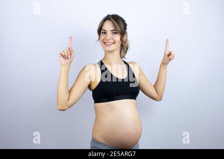 Jeune sportswoman enceinte portant des vêtements de sport sur fond blanc souriant, regardant la caméra et pointant vers le haut avec les doigts et les bras relevés Banque D'Images