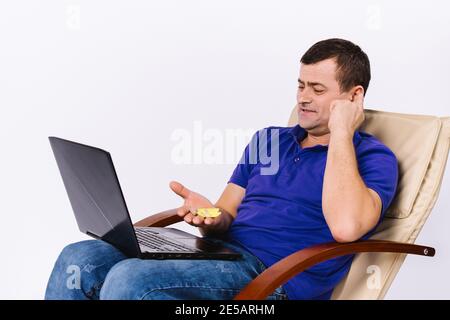 Un homme sourd âgé dans une chaise en cuir avec un ordinateur portable et un sourire montre une prothèse auditive à la caméra. Blog de motivation de santé en ligne. Arrière-plan blanc. Banque D'Images