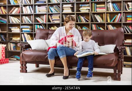 Femme caucasienne tenant sa petite fille et assise à côté de son fils sur un canapé à la bibliothèque à la maison. Mère et garçon lisant un livre intéressant. Banque D'Images