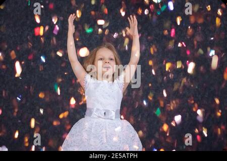 Belle petite fille dans une robe blanche avec des mains levées sous des confettis lumineux. Banque D'Images