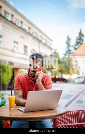 Smliling homme parlant sur un téléphone tout en étant assis devant un ordinateur portable dans le café Banque D'Images