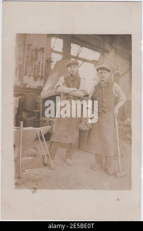 Portrait de deux forgerons édouardiens debout dans leur forge, l'un tenant un grand marteau. L'enclume est à gauche et les rangées d'outils sont alignées le long du mur. Banque D'Images