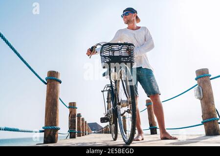 Portrait d'un homme heureux souriant pieds nus vêtu de vêtements d'été légers et de lunettes de soleil à vélo sur la jetée en bois. Des vacances imprudentes Banque D'Images