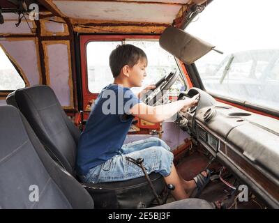 Un garçon de 11 ans joue comme conducteur derrière le volant d'une vieille voiture abandonnée. Banque D'Images