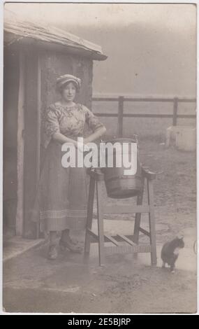 Femme debout dans une cour de ferme à côté d'une urne de beurre, le chat de ferme est à côté d'elle, 1900s / 1910s Banque D'Images