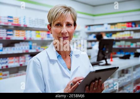 Un pharmacien d'âge moyen expérimenté fait défiler une tablette numérique pour regarder un appareil photo debout en pharmacie Banque D'Images