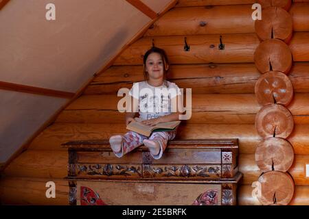 une jeune fille espiègle et espiègle est assise sur une vieille commode en bois avec ses jambes en haut et un livre ouvert sur ses genoux dans la chambre d'une maison rustique en rondins Banque D'Images