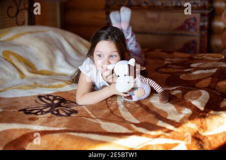 Une jeune fille gaie et sortie tient une vache à jouets et se trouve un matin ensoleillé sur un lit dans la chambre d'une maison rustique en rondins Banque D'Images