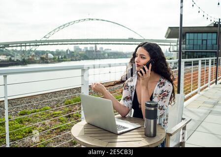 Femme assise sur le toit avec un ordinateur portable parlant au téléphone Banque D'Images