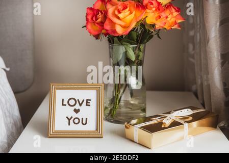 Boîte cadeau dorée avec ruban, un bouquet de fleurs roses dans un vase et un cadre avec Amour vous message sur la table de chevet contre un mur gris. Saint-Valentin Banque D'Images