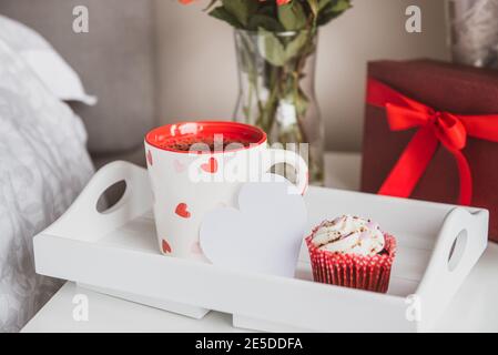 Plateau avec une tasse de thé chaud, un cupcake, une carte vierge en forme de coeur, une boîte cadeau et un bouquet de fleurs dans un vase sur la table de chevet. Saint Valentin Banque D'Images