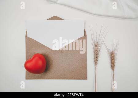 Le papier blanc vierge est placé sur une enveloppe de papier marron ouverte avec un coeur rouge et une fleur d'orge sèche et un tissu blanc sur fond blanc. Saint Valentin con Banque D'Images