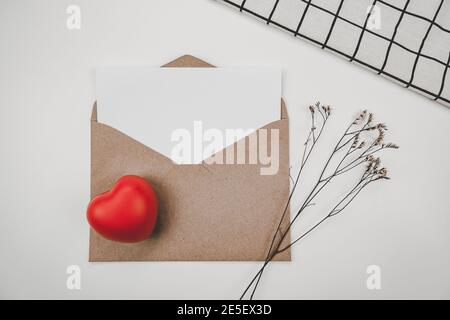 Le papier blanc vierge est placé sur une enveloppe de papier marron ouverte avec un cœur rouge et une fleur sèche de Limonium et un chiffon blanc sur fond blanc. Saint-Valentin c Banque D'Images