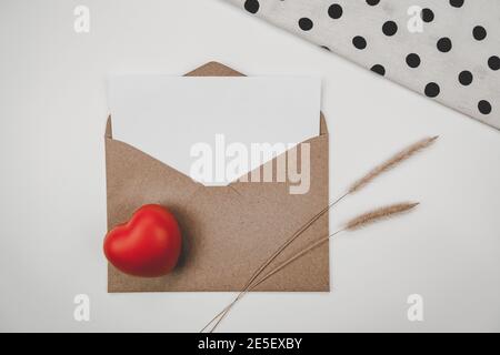Le papier blanc vierge est placé sur une enveloppe de papier brun ouvert avec un cœur rouge, une fleur sèche de queue d'éponge, un tissu blanc sur fond blanc. Saint Valentin Banque D'Images