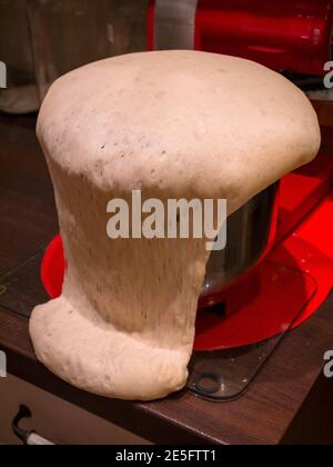 La pâte déborde d'un bol qui repose sur un tapis de verre sur une table en bois. En arrière-plan se trouve le robot de cuisine et le bloc rouge. Banque D'Images