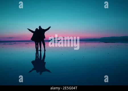 Style pop art bleu et rose silhouette de couple heureux ouvrant les bras sur la surface d'inondation des appartements de sel d'Uyuni au crépuscule, Bolivie Banque D'Images