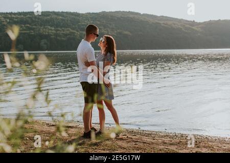 Portrait d'un couple amoureux se câliner sur la plage au coucher du soleil. Pleine longueur de jeune femme et d'homme qui embrasse le lac pendant la chaude soirée d'été. Banque D'Images