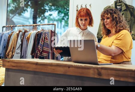 Deux femmes travaillant dans un magasin de vêtements à la recherche d'un ordinateur portable au comptoir. Partenaires travaillant ensemble dans une boutique de vêtements. Banque D'Images