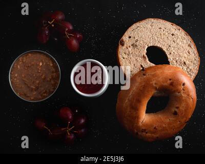 Un bagel à la cannelle et à la raisins secs avec une marmite de confiture, de beurre d'arachide et de raisins rouges sur une surface noire parsemée de sucre Banque D'Images