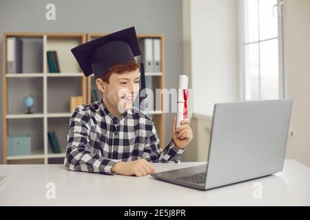 Garçon diplômé assis à un bureau dans un chapeau de troisième cycle et tenant un rouleau de diplôme devant un ordinateur portable. Banque D'Images