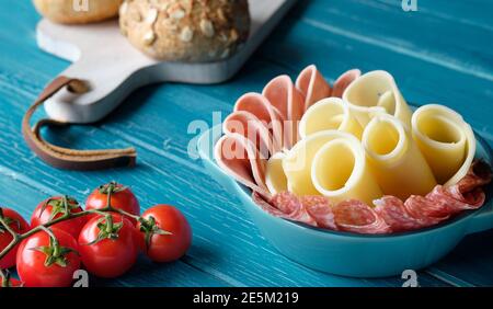 Salami italien, saucisse et fromage dans un bol en céramique. Faire une brindille avec des tomates cerises. Planche à découper avec pains à grains entiers.planches en bois turquoise. Banque D'Images