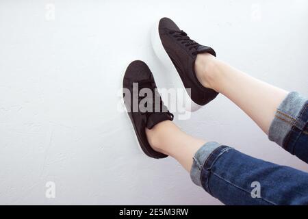 Pieds femelles vus d'en haut. Selfie pour toute utilisation. Pieds et jambes en baskets et jeans noirs. Pied levé sur un mur blanc. Avec espace de copie. Pieds à l'extérieur Banque D'Images