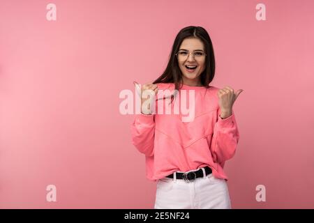 Photo d'une jeune femme attrayante en lunettes sourit et donne des pouces vers le haut. Porte un t-shirt décontracté rose pantalon blanc isolé couleur rose fond Banque D'Images
