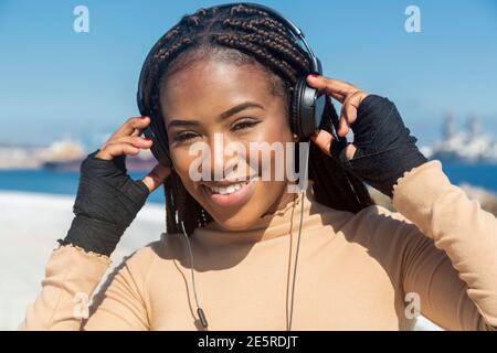 Portrait d'une jeune femme afro-américaine souriante avec casque regardant l'appareil photo, avec fond de mer et de ciel, promenade. Banque D'Images