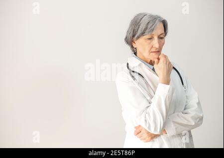 Portrait d'une femme mature senior médecin debout avec un stéthoscope, posant sur fond gris, regardant vers le bas et pensant, qualifié et expérimenté Banque D'Images