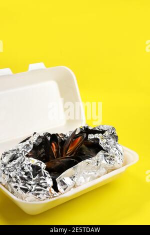 Livraison de nourriture dans un emballage en plastique à votre maison. Moules dans une sauce en papier d'aluminium et contenant en plastique sur fond jaune. Banque D'Images