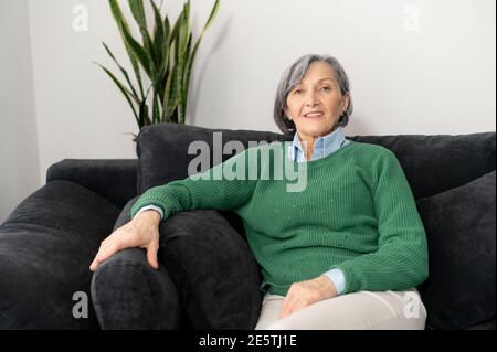 Belle femme sénior mûre grise dans un pull vert décontracté est penchée sur un canapé dans une position confortable, prenant une pause de travail, milieu Banque D'Images