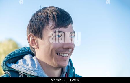 Vue latérale de jeune adulte caucasien cheveux foncés homme souriant à l'extérieur, le jour ensoleillé de l'automne ou du printemps contre le bleu ciel Banque D'Images