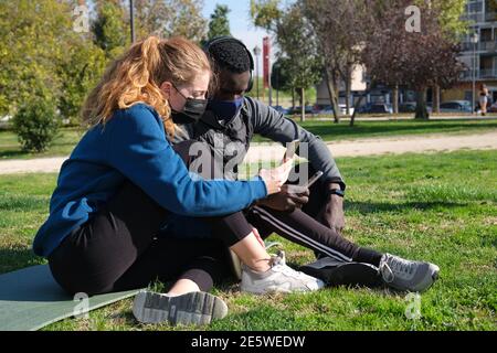Homme noir africain et femme caucasienne portant des masques de visage vérifiant le smartphone après avoir fait de l'exercice dans un parc avec un tapis. Fitness et sport outd Banque D'Images