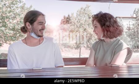 De jeunes amis portant des masques de visage ayant une conversation tout en étant assis sur un banc en bois dans le parc. Rencontrer des amis et s'amuser pendant une pandémie Banque D'Images