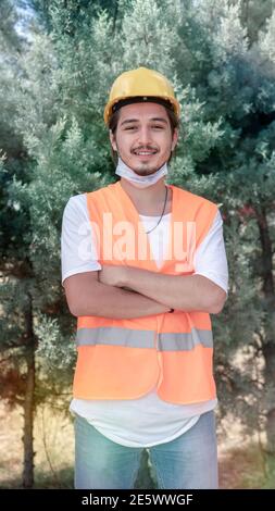 Portrait d'un jeune ouvrier de construction avec un casque jaune et un masque facial debout avec les bras croisés et regardant la caméra sur le chantier. Banque D'Images
