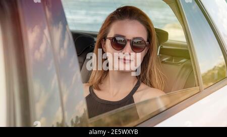 Bonne jeune belle femme en lunettes de soleil assis dans la voiture regardant l'appareil photo. Banque D'Images