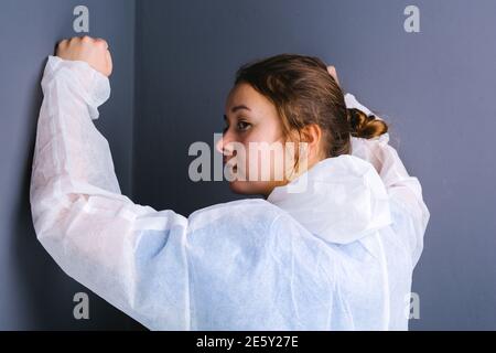Photo d'une femme médecin fatiguée qui a posé ses poings contre un mur gris et regarde loin. Photo de derrière. Banque D'Images