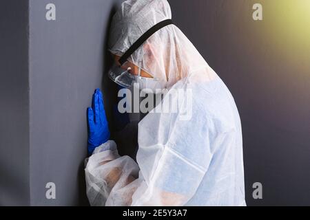 Un médecin fatigué en équipement médical de protection et des gants en latex jetables bleus a posé sa tête contre le mur gris de l'isolement. Banque D'Images