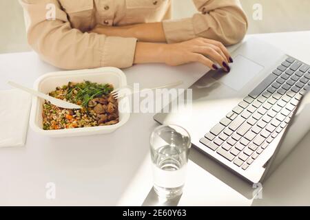 Femme d'affaires assise à un bureau, prenant un déjeuner à emporter, de l'eau potable et utilisant un ordinateur portable Banque D'Images