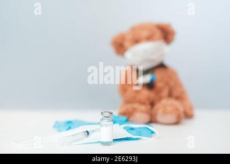 Gants médicaux, seringue et flacon de vaccin contre le coronavirus Covid-19 en premier plan, l'ours en peluche dans un masque de protection se trouve en arrière-plan Banque D'Images