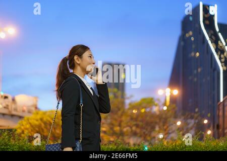 Femme de direction asiatique travaillant à l'aide d'un téléphone mobile dans la rue avec des immeubles de bureaux en arrière-plan la nuit à Bangkok, en Thaïlande. Banque D'Images